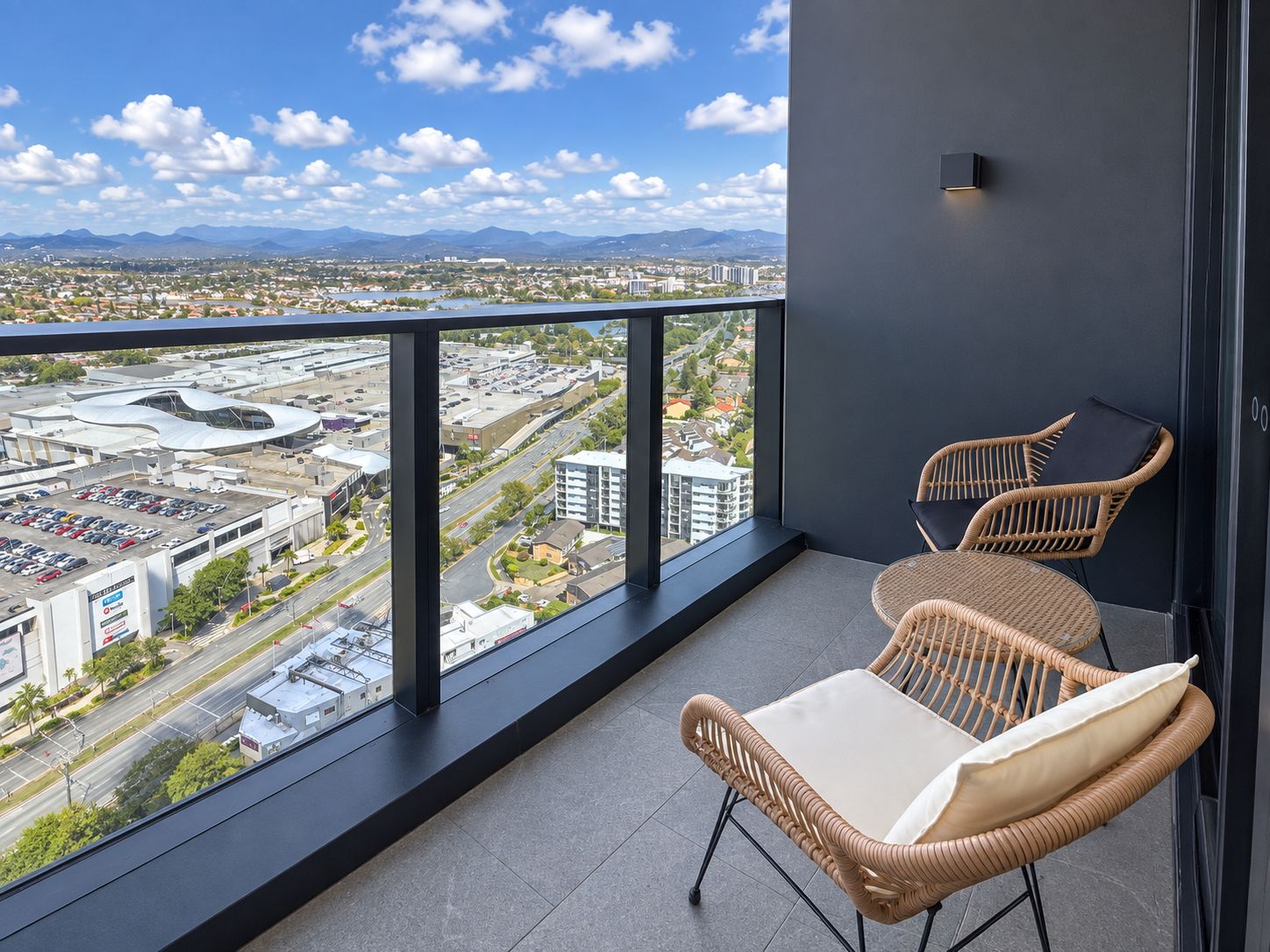A balcony with wicker chairs and a view of a cityscape, including a large building and mountains in the distance.