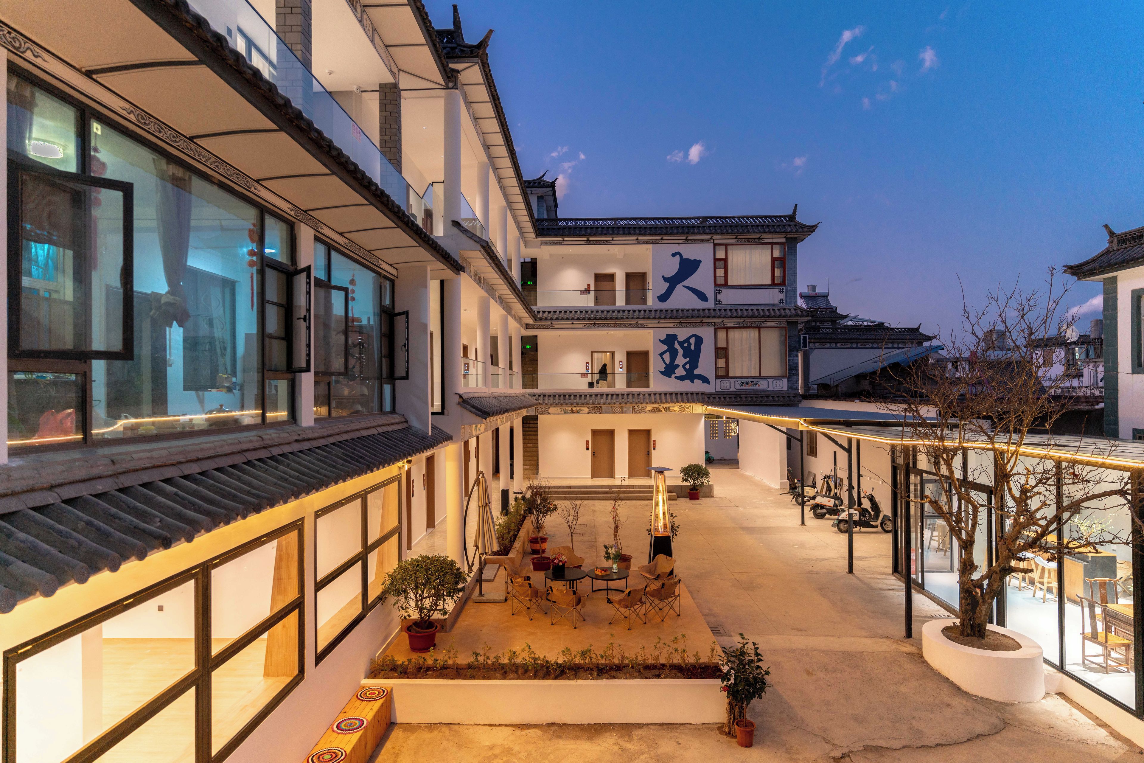 Un edificio tradicional con patio, que presenta un letrero grande con caracteres chinos, un área de descanso con sillas y mesas, y un espacio para estacionar scooters.