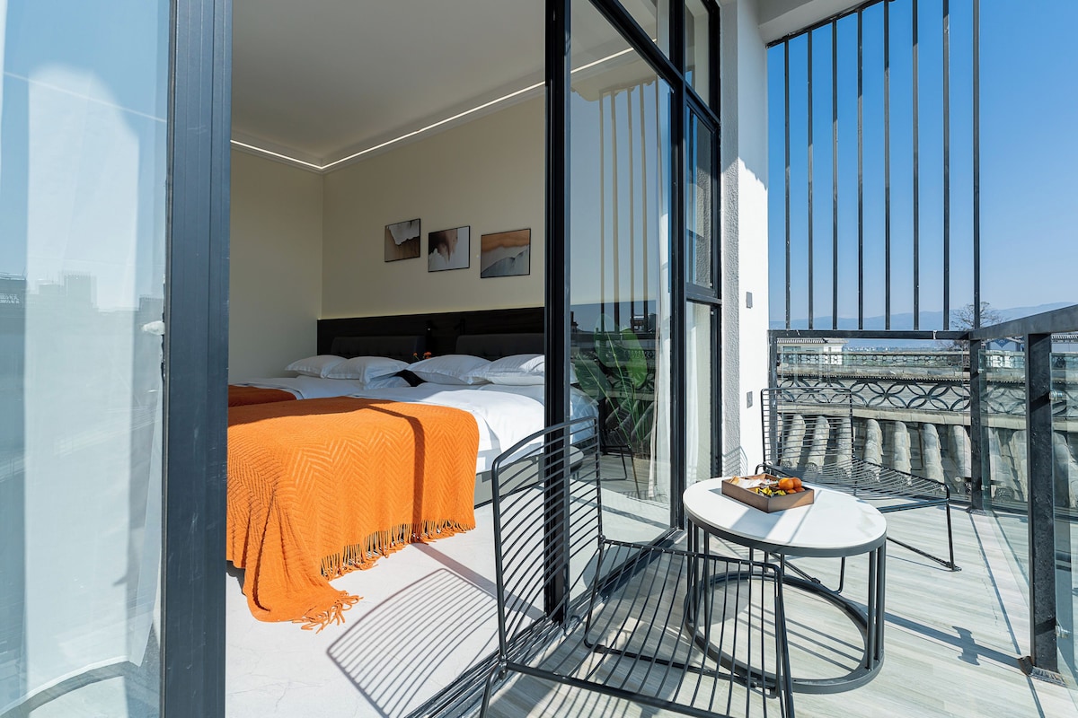 Habitació d'hotel moderna amb un llit, una taula petita amb una safata de fruita i un balcó amb vista a la ciutat.