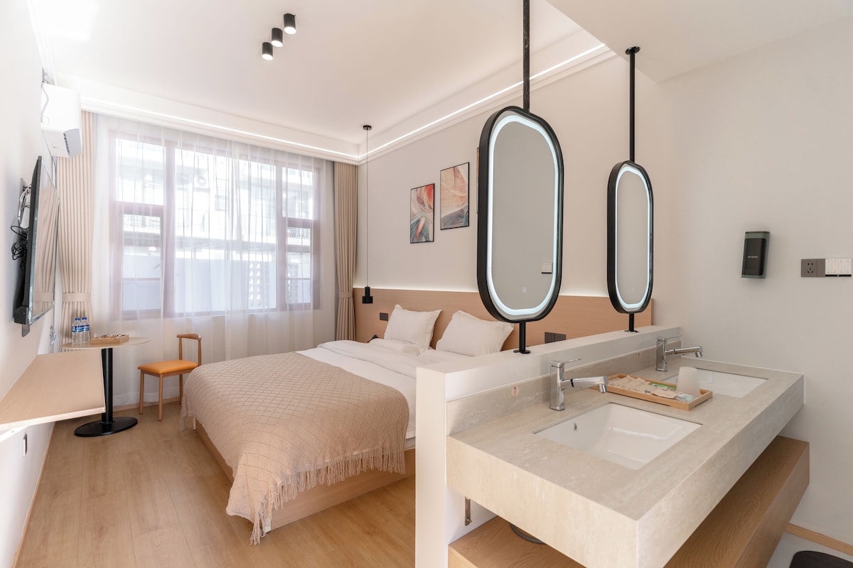 Habitació d'hotel moderna amb llit, taules de nit, escriptori, cadira, finestra gran amb cortines, televisió penjada a la paret, dos miralls penjats i bany amb doble mànega i barra de tovalloles.