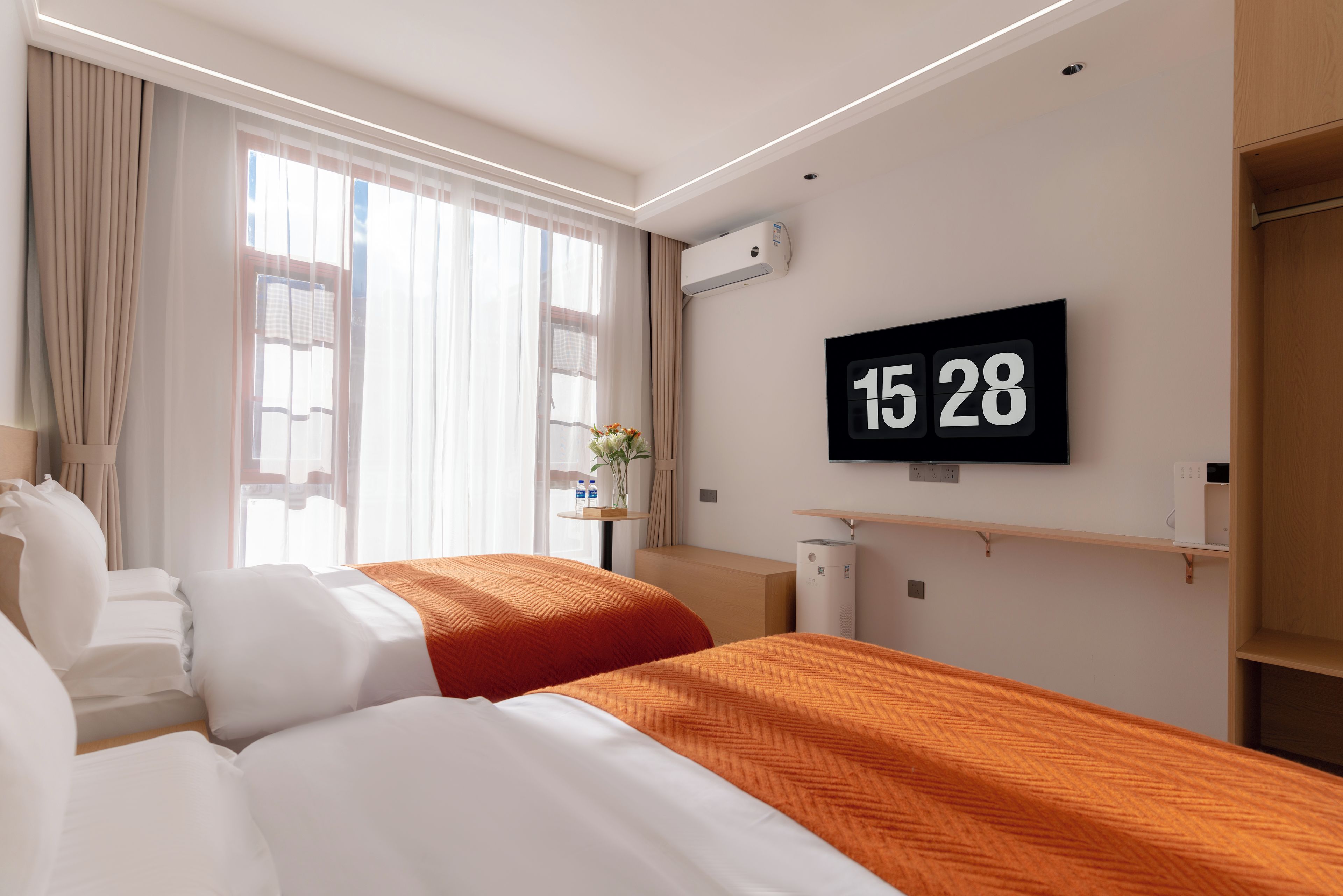 Une chambre d’hôtel avec deux lits, un téléviseur fixé au mur et une horloge numérique indiquant l’heure.