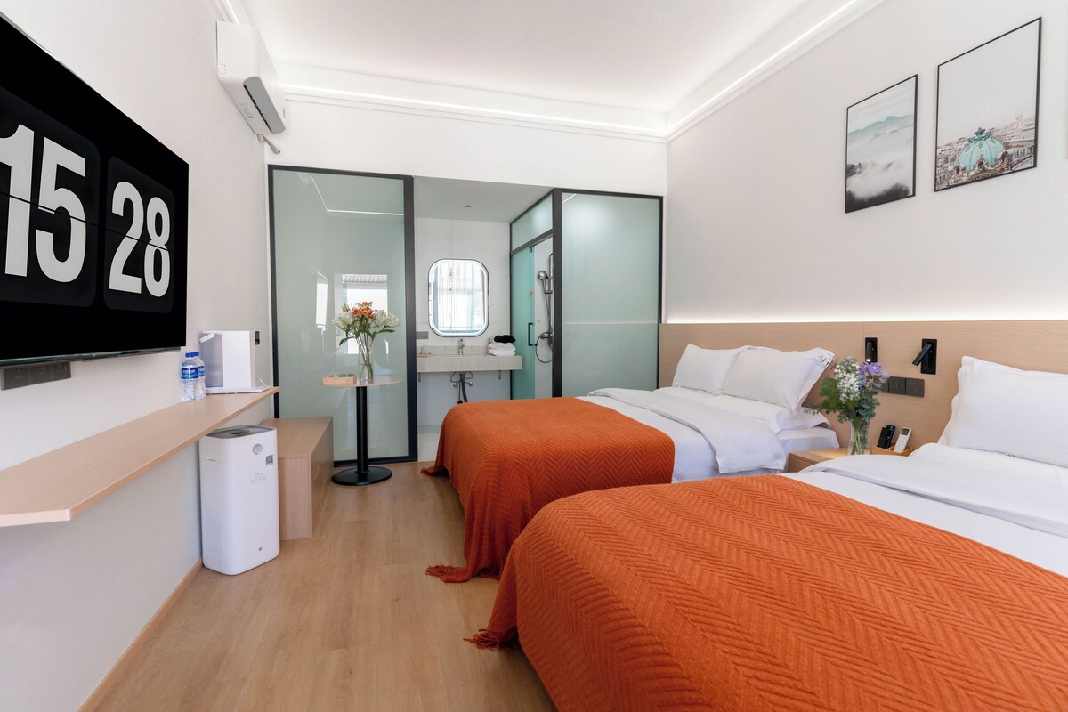 Habitació d'hotel amb dos llits, una taula de treball, un televisor i un bany visible a través d'una porta de vidre.