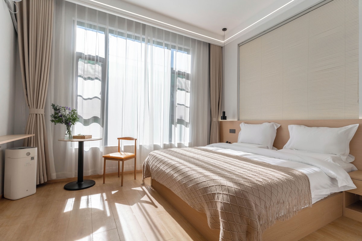 Habitació d'hotel amb un llit, una cadira, una taula petita i una finestra amb cortines.