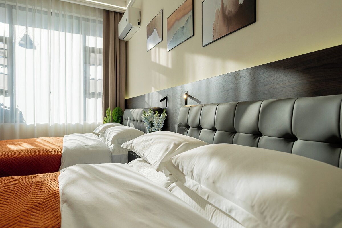 Una habitació d'hotel amb dos llits, una finestra gran amb cortines i aire condicionat fixat a la paret.