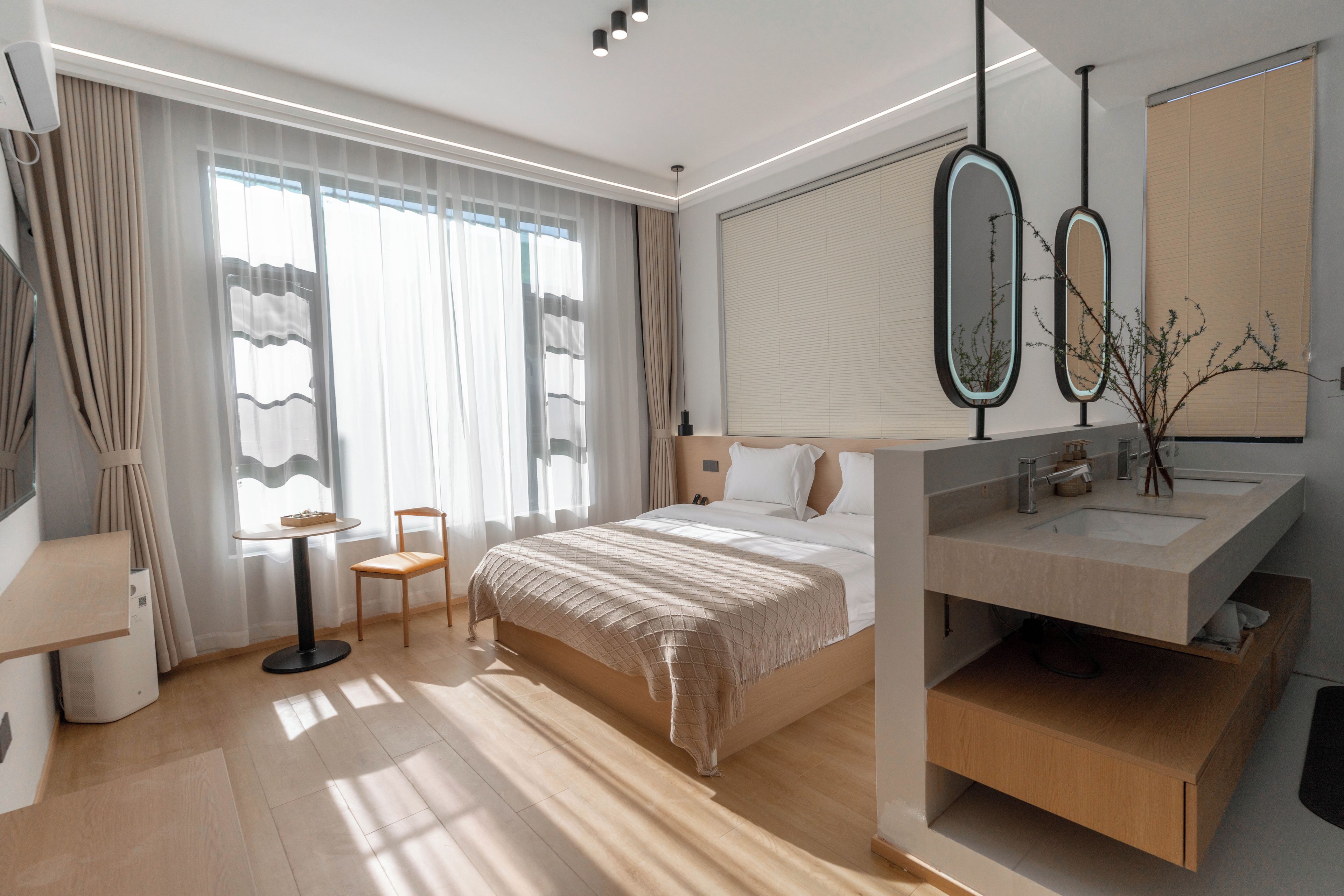A modern hotel room with a large bed, a bedside table, a chair, a desk, a large window with curtains, a bathroom with a sink and mirror, and a plant.
