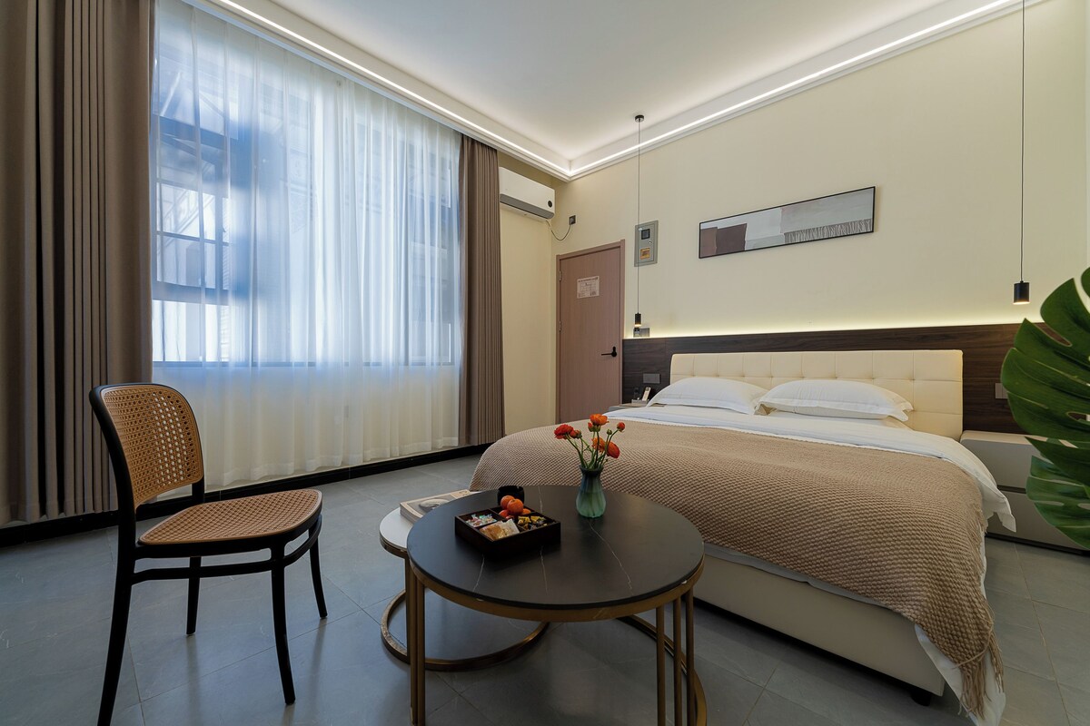 Una habitació d'hotel amb un llit, una cadira, una taula i una finestra amb cortines.