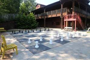 A large outdoor chessboard set up on a concrete surface with a wooden building in the background.