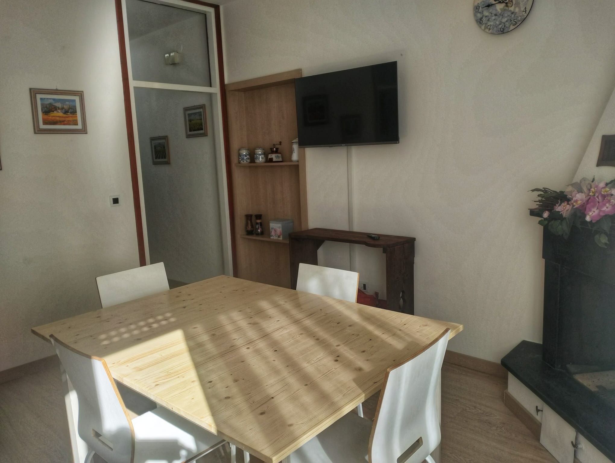 Uma sala de jantar com mesa de madeira e cadeiras brancas, televisão fixada na parede e um pequeno móvel decorativo.