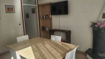 Uma sala de jantar com mesa de madeira e cadeiras brancas, televisão fixada na parede e um pequeno móvel decorativo.