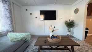 Een woonkamer met een bank, een bijzettafeltje, een flatscreen televisie aan de muur, een potplant en een decoratieve vaas.