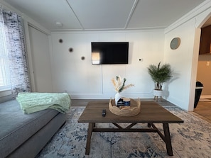 En stue med sofa, sofabord, fladsk ærms-tv monteret på væggen, en potteplante og en dekorativ vase.
