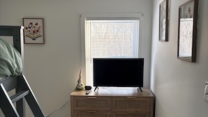 Een slaapkamer met een stapelbed, een houten dressoir, een flatscreen televisie en een raam met jaloezieën.