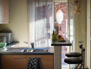 Een keuken met een spoelbak, een raam met uitzicht op een bakstenen gebouw, een barkruk en een hangende lamp.