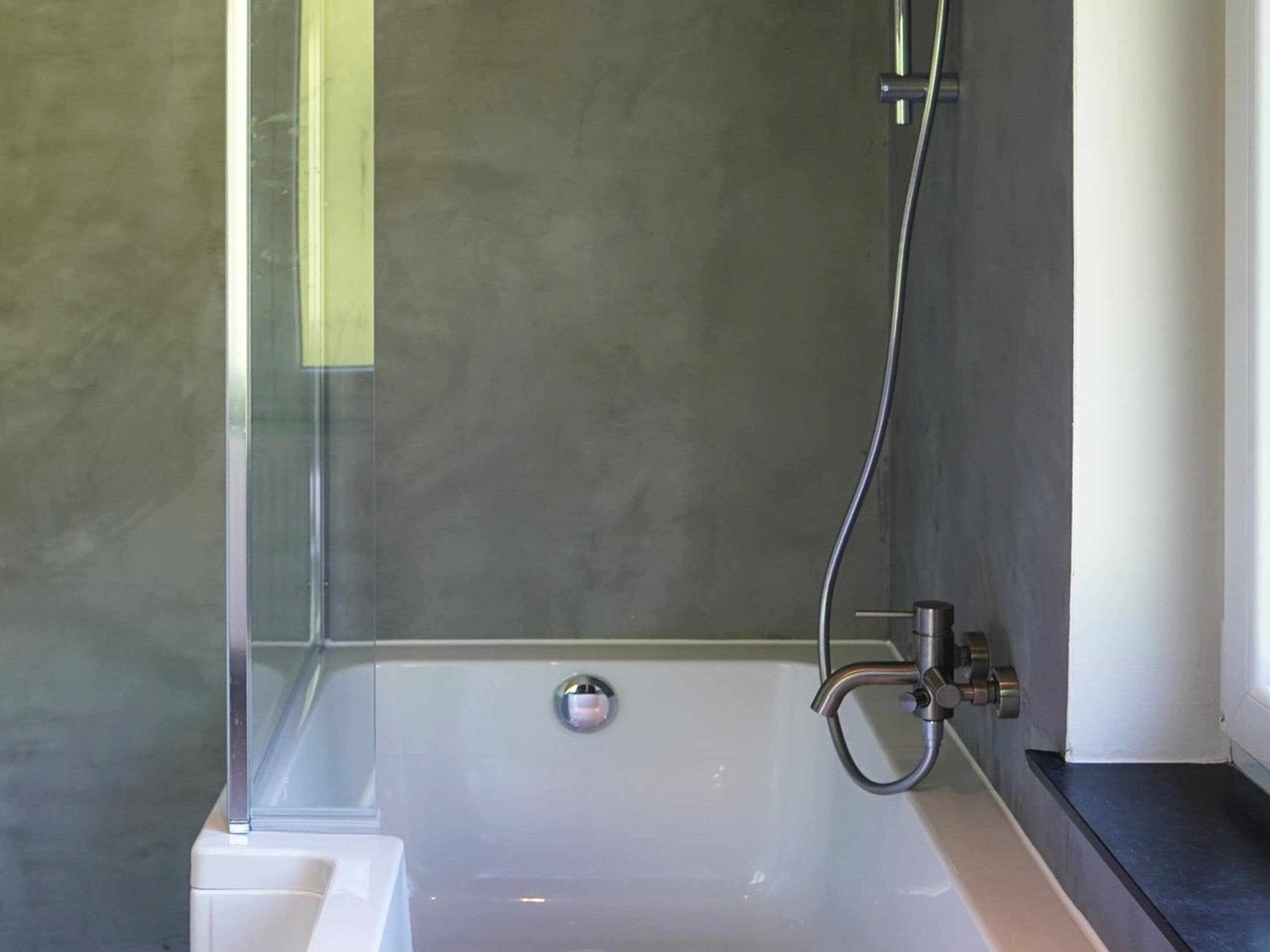 Een moderne badkamer met een douche met glazen wand, een wit bad en een grijze betegelde muur.