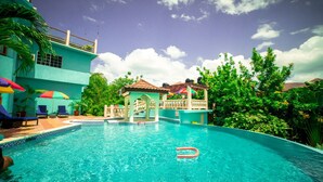 En swimmingpool med klart, blåt vand, omgivet af frodig vegetation og en bygning med flere niveauer og et flisegulv.