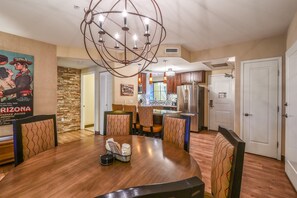 A dining area with a round table, chairs, a chandelier, a kitchen in the background, and a poster on the wall.