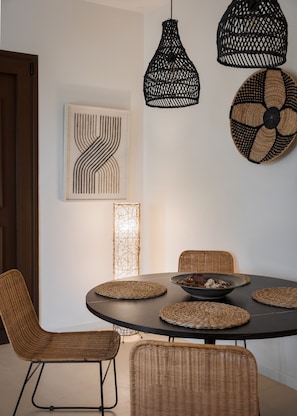 A dining area with a round table, woven chairs, and a black pendant light. - Casa Smaragdi Aegina (Perdika)