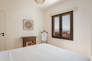A bedroom with a bed, a wooden desk, a hanging towel, a framed wall decoration, and a window with a view. - Casa Smaragdi Aegina (Perdika)