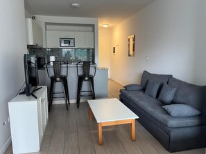 Ein modernes Wohnzimmer mit einem grauen Sofa, einem weißen Couchtisch und einer Küchenzeile inklusive Mikrowelle und zwei Stühlen.