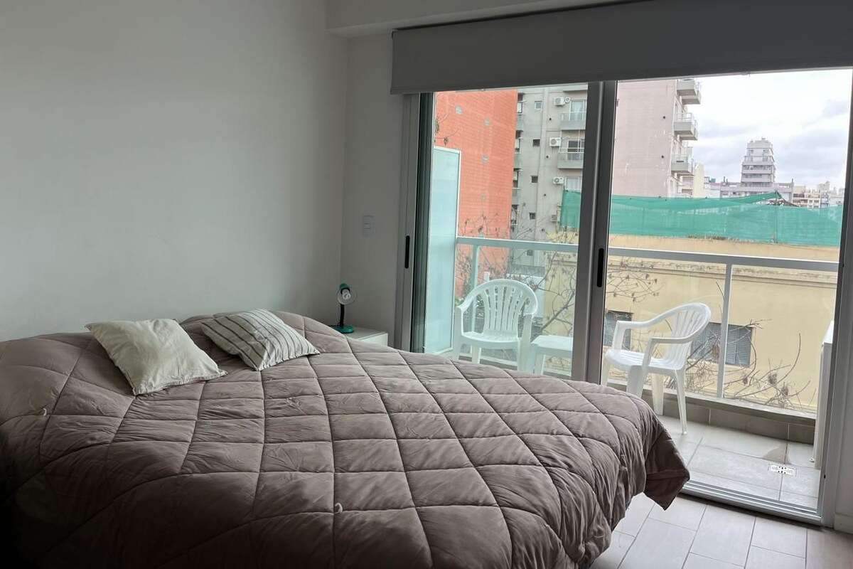 Ein Schlafzimmer mit einem großen Bett, einem Nachttisch, einem Ventilator und Blick auf einen Balkon mit Stühlen und Gebäuden.