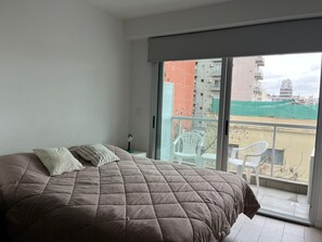 Ein Schlafzimmer mit einem großen Bett, einem Nachttisch, einem Ventilator und Blick auf einen Balkon mit Stühlen und Gebäuden.