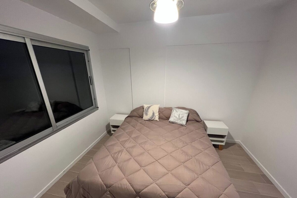 Ein Schlafzimmer mit einem großen Bett, zwei Nachttischen, einem Fenster und einer Deckenleuchte.