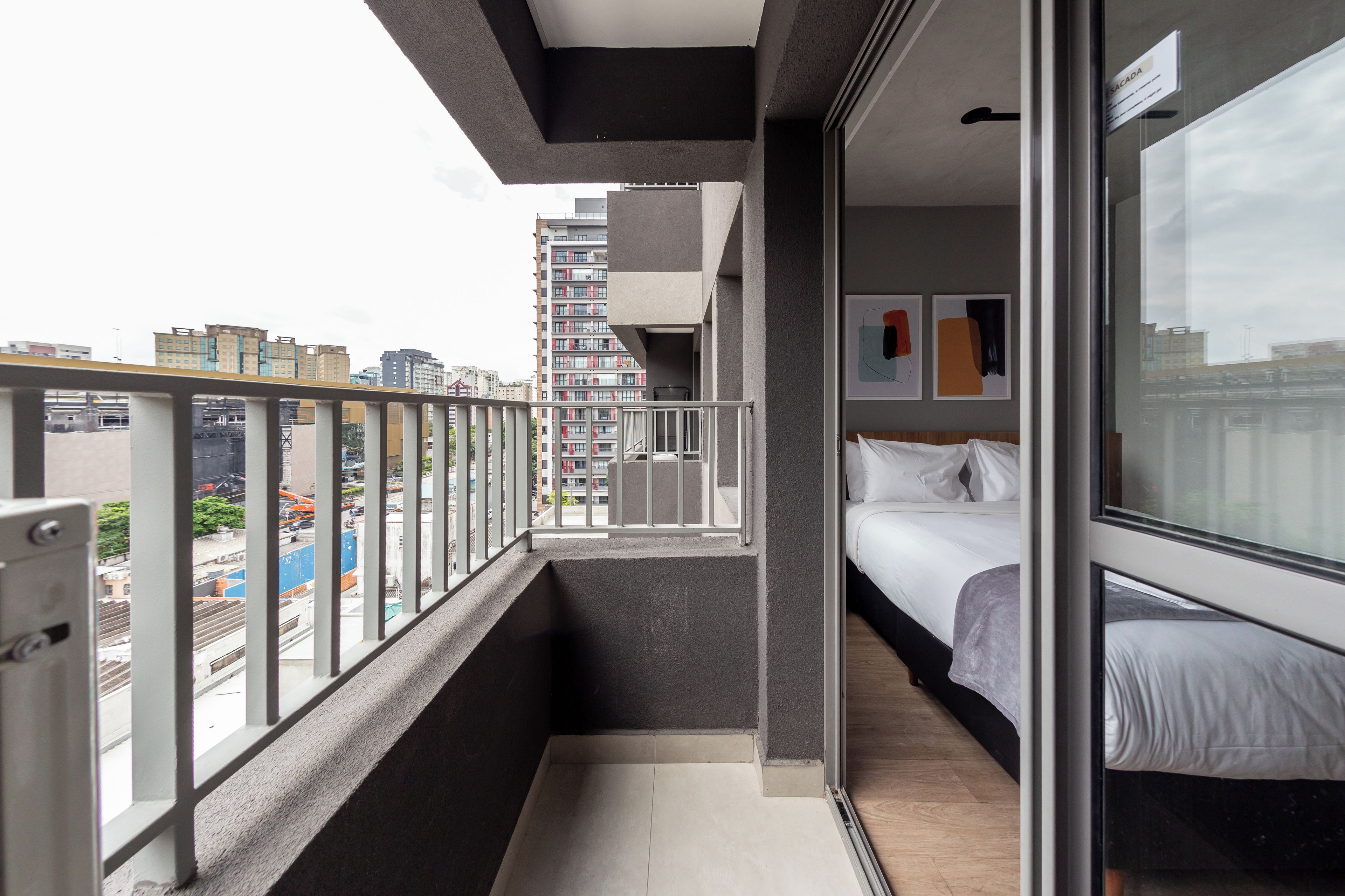 A narrow balcony with a city view, a bed with white bedding, and a door leading to a room with a window.