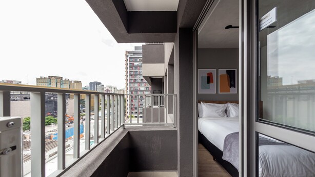 Varanda estreita com vista para a cidade, cama com roupa de cama branca e porta que dá para um quarto com janela.