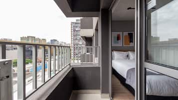 A narrow balcony with a city view, a bed with white bedding, and a door leading to a room with a window.