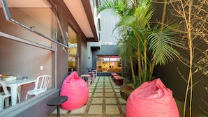 Um corredor externo estreito com chão de tijolos de pedra, dois bean bags cor-de-rosa e uma área de estar com mesas e cadeiras.