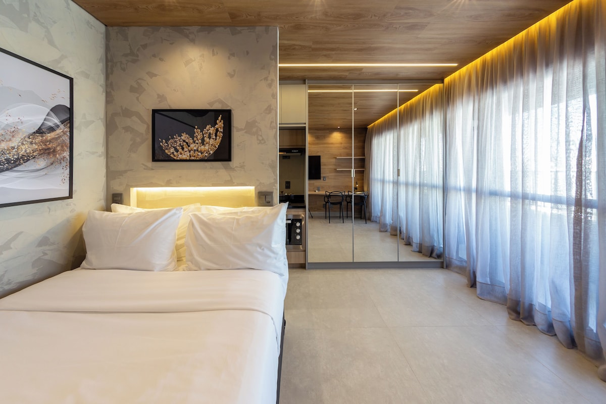 A modern hotel room with a bed, a dining area, and a view through sheer curtains.