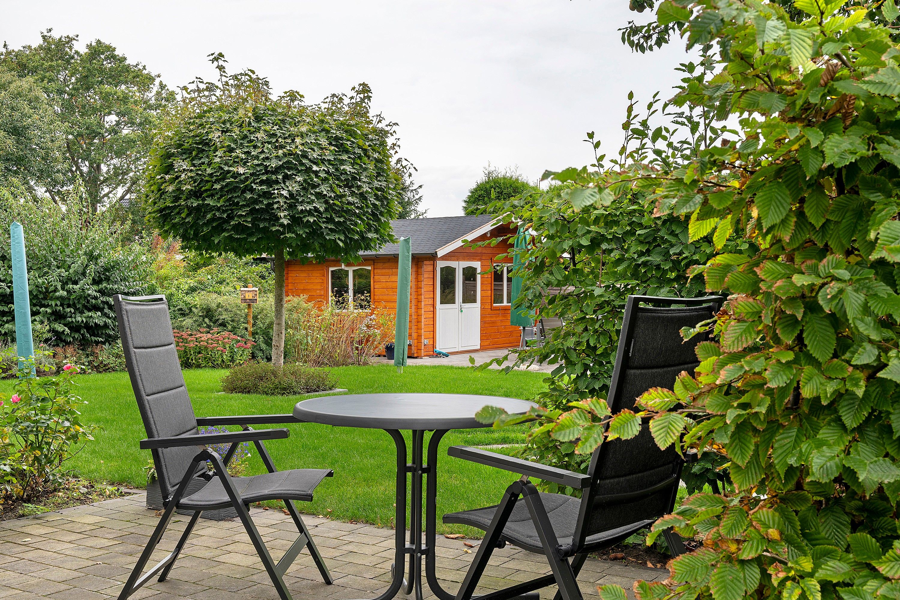An outdoor seating area with a table and chairs, a small wooden structure in the background, and lush greenery.