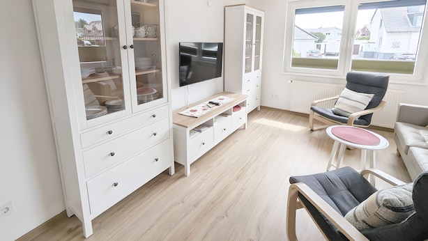 Ein Wohnzimmer mit einem weißen Schrank, einem Fernseher, einem Sessel und Blick ins Freie.