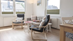 Ein modernes Wohnzimmer mit einer Couch, zwei Schaukelstühlen, einem kleinen Tisch und großen Fenstern mit Blick auf Häuser.