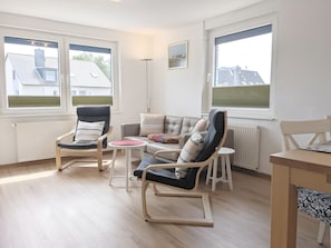 Uma sala de estar moderna com um sofá, duas poltronas de balanço, uma mesa pequena e janelas amplas que oferecem vista para casas.