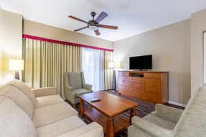 A hotel room with a sofa, armchair, wooden coffee table, television, and a ceiling fan. - Pompano Beach | Family Golf Vacation | Pool + Golf | 2BR (Pompano Beach)