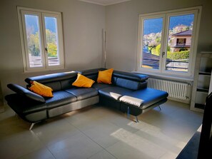 A modern living room with a sectional sofa, two windows with a view of a residential area, and a radiator. - Charming, recently renovated apartment with a balcony overlooking the river (Cannobio)