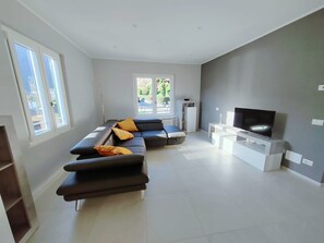 Ein modernes Wohnzimmer mit einer Eckcouch, einem Flachbildfernseher und Blick auf einen Garten durch große Fenster.