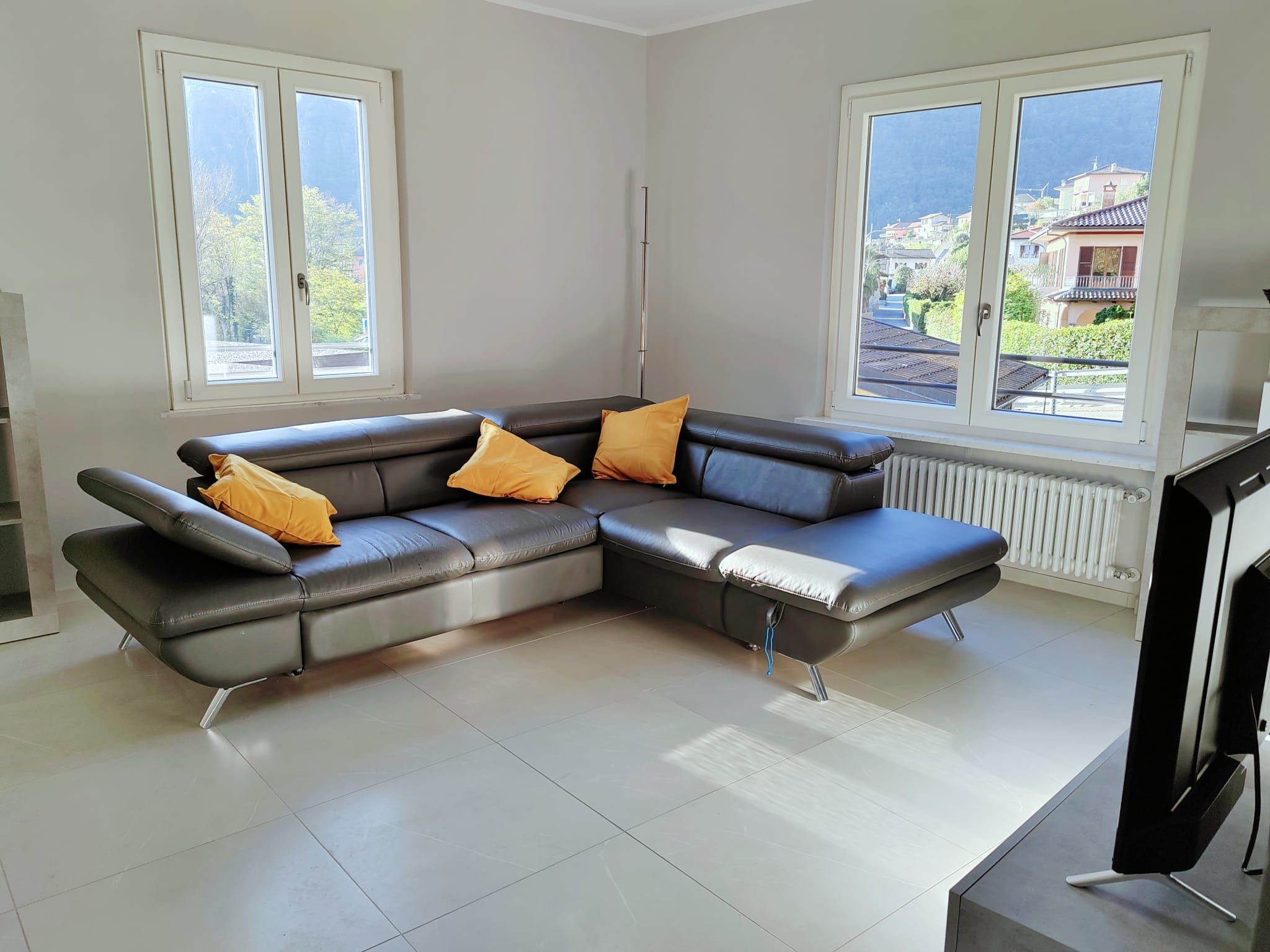 Ein modernes Wohnzimmer mit einem Ecksofa, zwei Fenstern und Blick auf eine Wohngegend.