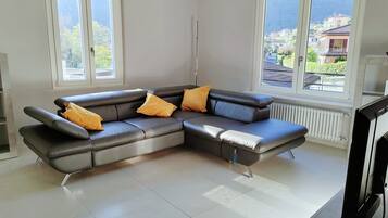 Ein modernes Wohnzimmer mit einem Ecksofa, zwei Fenstern und Blick auf eine Wohngegend.