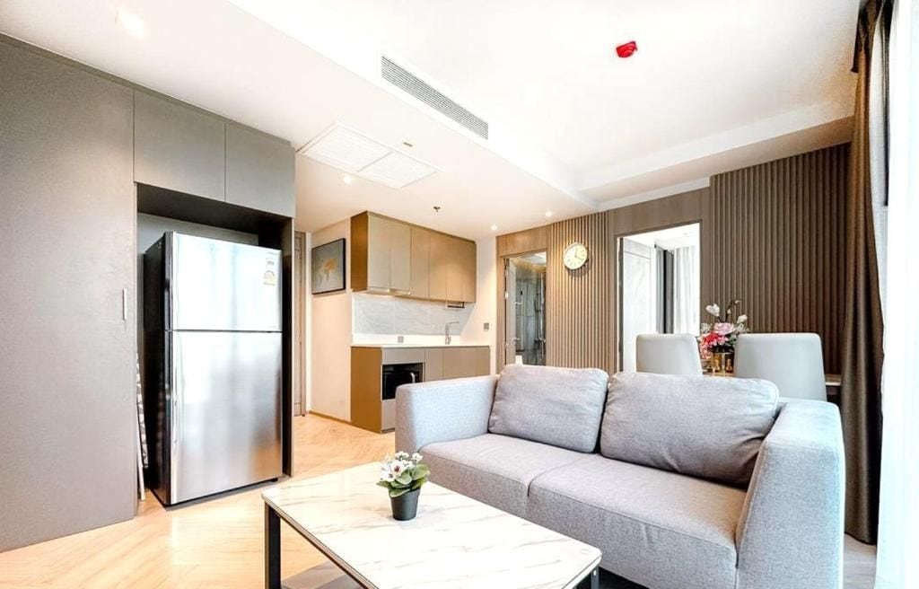A modern living room with a grey sofa, a coffee table, and a kitchen area with a refrigerator and cabinets.