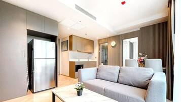 A modern living room with a grey sofa, a coffee table, and a kitchen area with a refrigerator and cabinets.