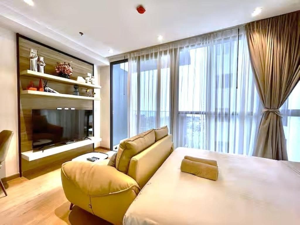 A modern hotel room with a large bed, a sofa, a TV mounted on the wall, and a view of the city through the window.