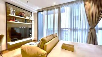 A modern hotel room with a large bed, a sofa, a TV mounted on the wall, and a view of the city through the window.