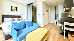 A hotel room with a bed, a blue sofa, a wooden table, a kitchenette, and a bathroom with a shower. - Andromeda Condominium Pattaya (Muang Pattaya)
