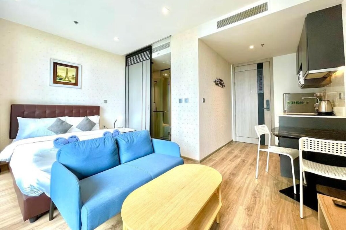 A hotel room with a bed, a blue sofa, a wooden table, a kitchenette, and a bathroom with a shower.