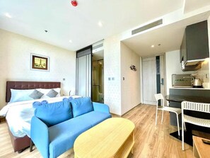 A hotel room with a bed, a blue sofa, a wooden table, a kitchenette, and a bathroom with a shower. - Andromeda Condominium Pattaya (Muang Pattaya)