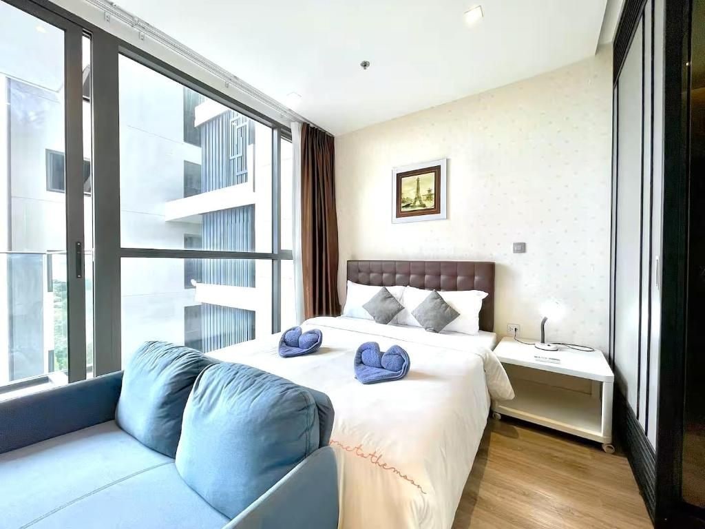 A hotel room with a large window, a bed with a white comforter, a blue sofa, and a picture of the Eiffel Tower on the wall.