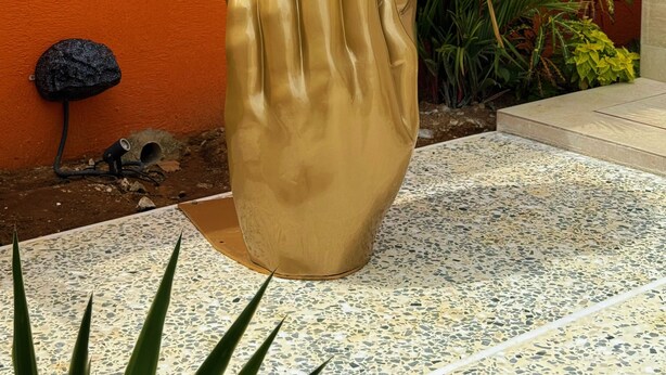 Eine Skulptur einer Hand in einem Garten mit orangefarbenen Wänden und Grünpflanzen.