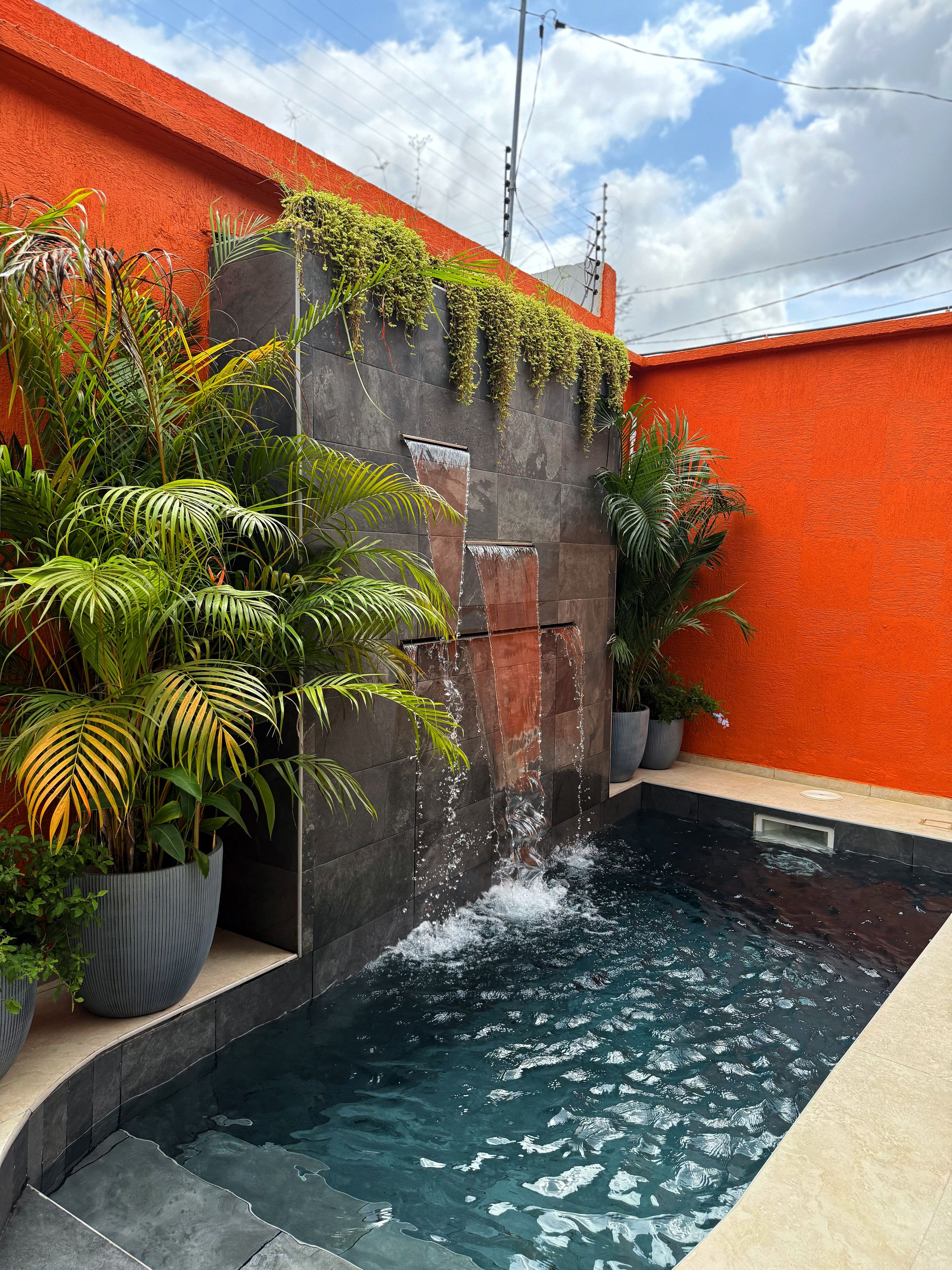 Une piscine avec de l’eau qui dévale le long d’un mur, entourée de plantes en pot et d’un mur orange.
